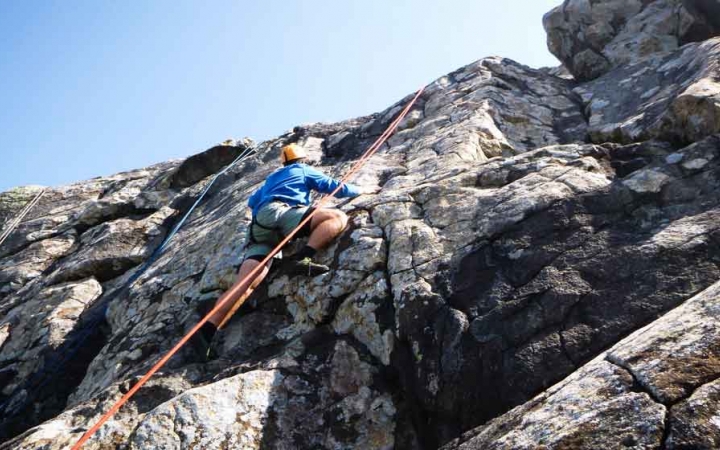 a person rock climbs a gray rock face under blue skies on an outward bound expedition in maine